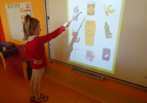 Dziewczynka stoi pod tablicą interaktywną i wskaźnikiem pokazuje puszkę metalową.